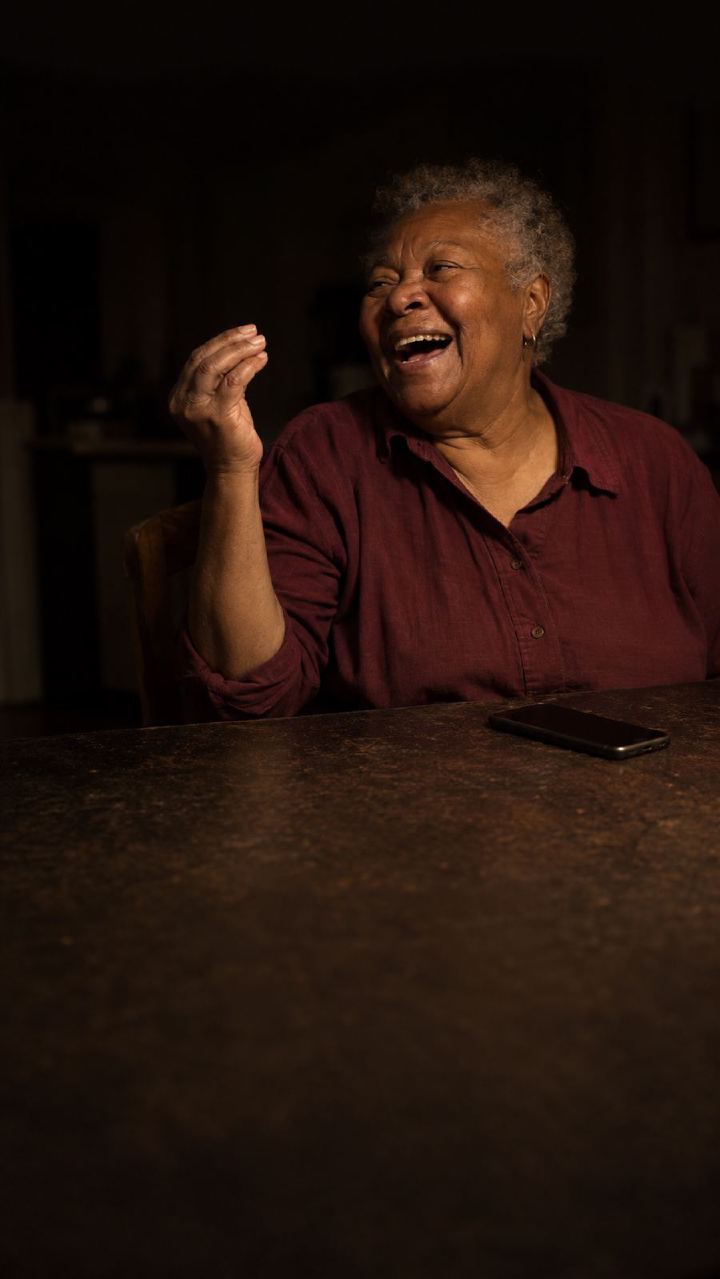 An older woman laughing mid-sentence at a kitchen table in lamplight, a phone recording in front of her